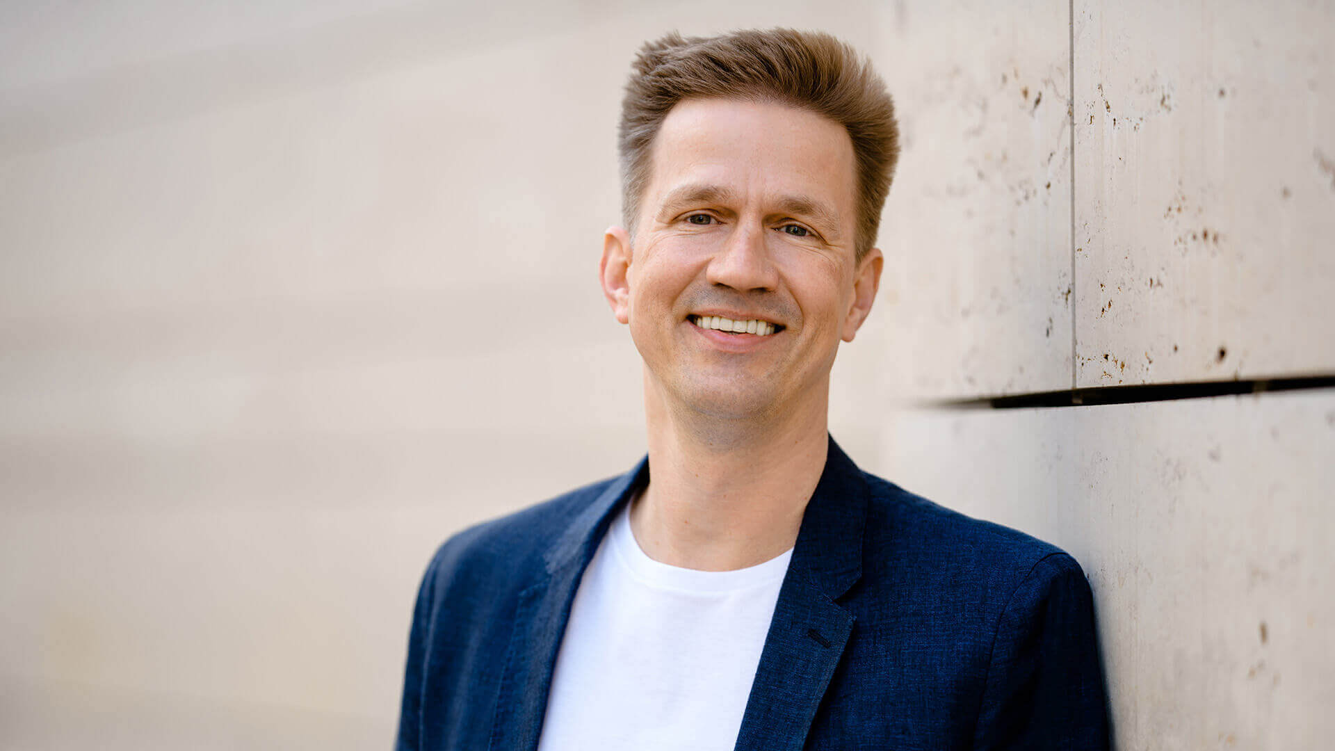 David Neumann in weißem T-Shirt und dunkelblauem Sakko, lächelnd an einer hellen Wand